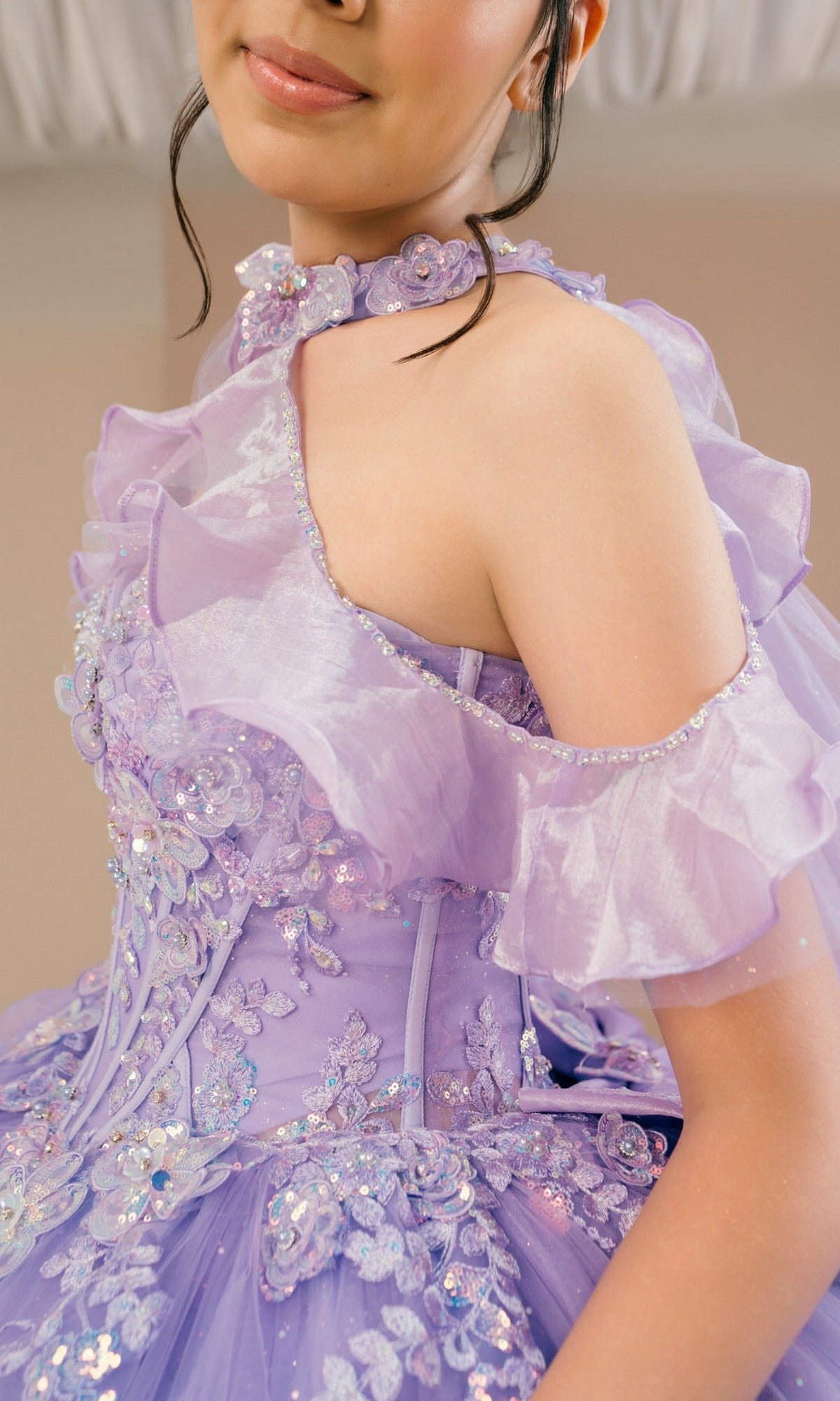 Wearing the Quinceanera Dress 8585JQ By Cinderella Couture, a lavender off-shoulder gown adorned with ruffled floral lace, appliqué, sequins, and a glitter tulle skirt, the person poses indoors.